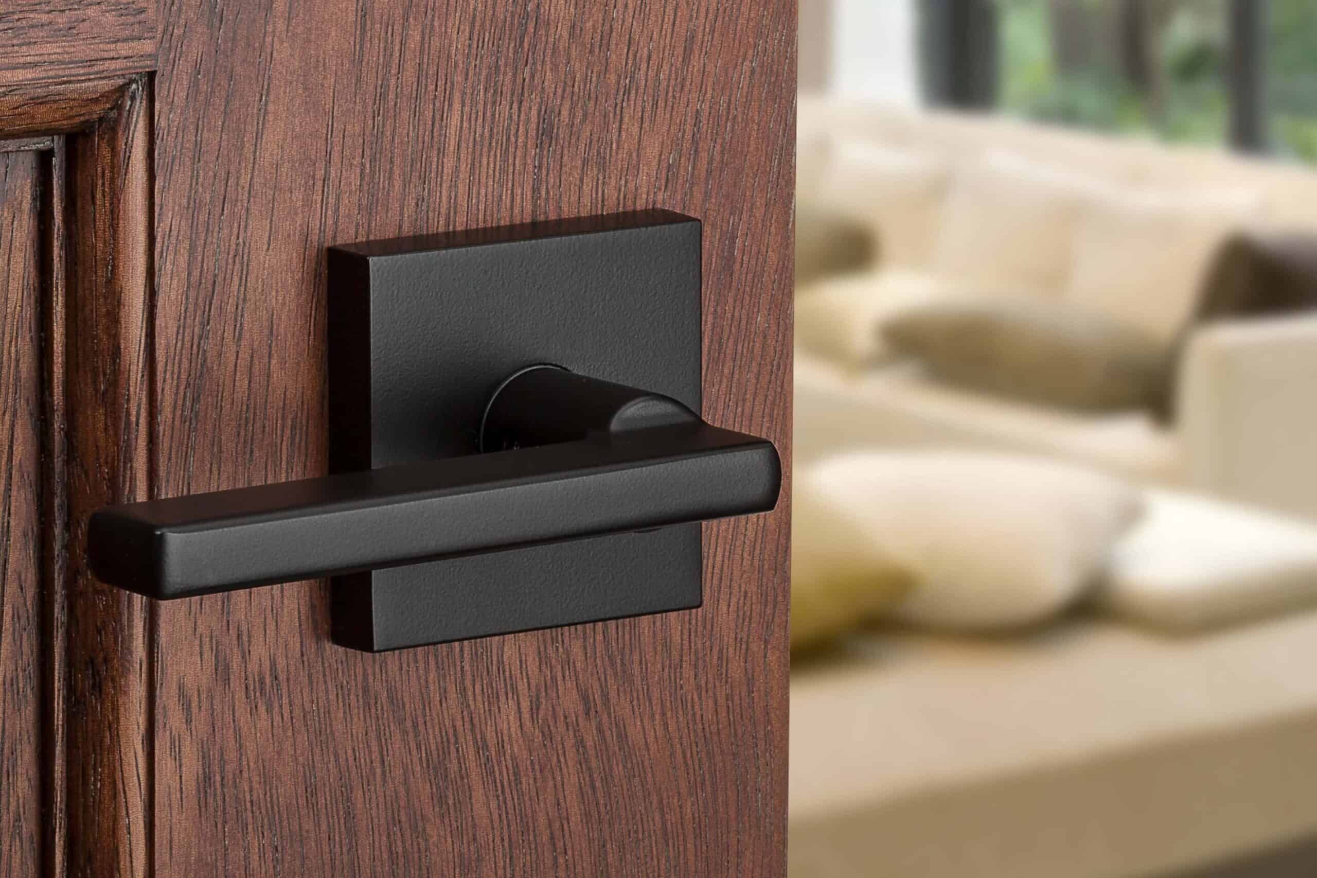 Close-up of sleek black door hardware on a wooden door, with a blurred view of a beige couch and pillows in the background.