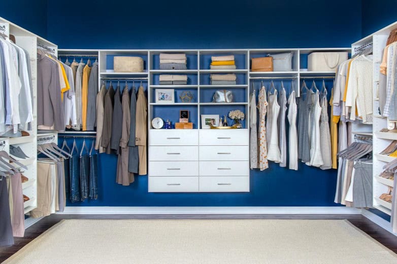 A neatly organized walk-in closet with various clothing items, purses, and accessories on shelves and hangers against a blue wall.