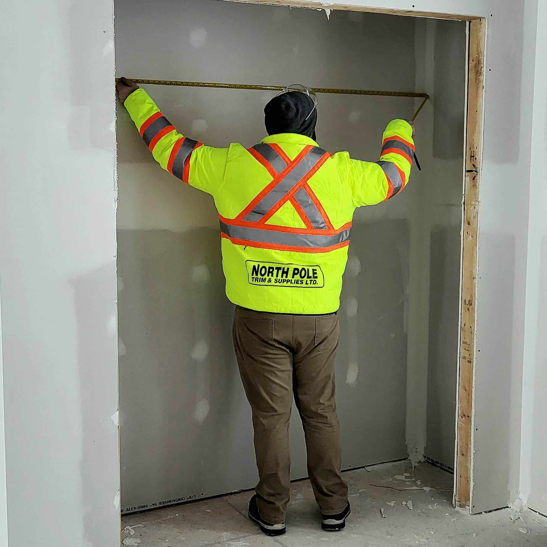 Person in a bright yellow safety jacket measures a doorway frame with a tape measure.