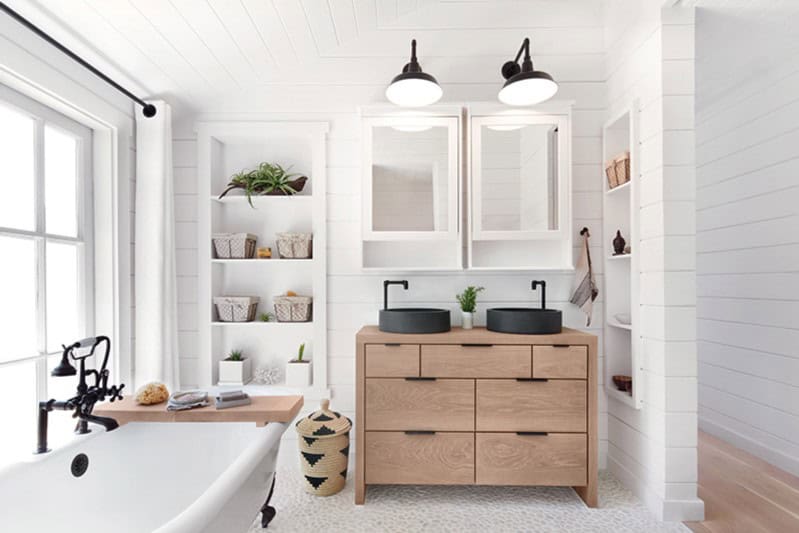 Modern bathroom with double vanity, black sinks, and wooden cabinets. Freestanding tub by a window, with shelves holding plants and baskets. White walls and ceiling with black light fixtures.